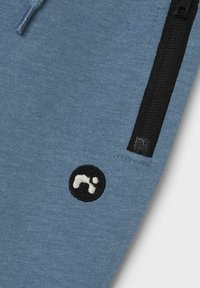 Zilā džemperi audums ar melnu rāvējslēdzēja kabatu un apļa formas melnu logotipu ar baltu simbolu. Tekstūra izskatās mīksta ar gludu virsmu.