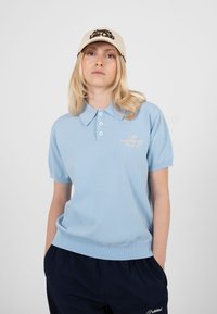 Femme aux cheveux blonds portant une casquette beige avec l'inscription « Always Late Club », un polo bleu clair et un pantalon foncé, debout devant un fond uni.