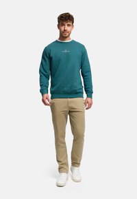 Bluza w kolorze morskim z okrągłym dekoltem i logo na środku, zestawiona z jasnobrązowymi spodniami i białymi tenisówkami. Materiał wygląda na miękki i teksturowany.