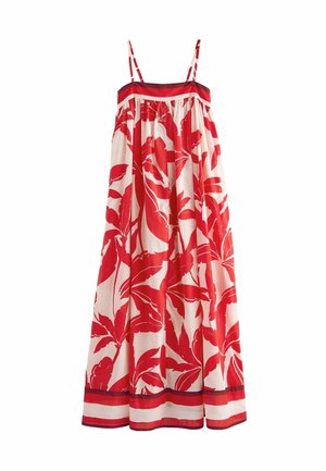 Donna che indossa un lungo abito senza spalline con motivi di foglie tropicali rosse, in piedi all'aperto su un pavimento in pietra, con infradito nere.