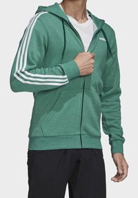 Sudadera verde con cremallera, hecha de un tejido suave, que presenta acentos en blanco con tres rayas en las mangas, capucha con cordones y bolsillos delanteros.