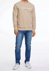 Felpa beige con la scritta "Copenhagen Lindbergh" stampata in bianco, abbinata a jeans blu e sneakers bianche, creando un look casual.