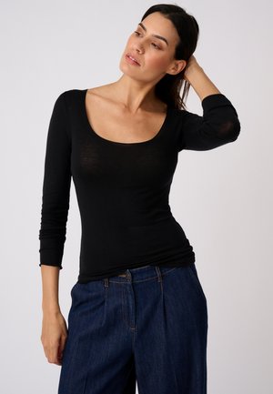 Donna con capelli scuri che indossa una maglia nera a maniche lunghe e jeans blu, posando con una mano dietro la testa su uno sfondo semplice.