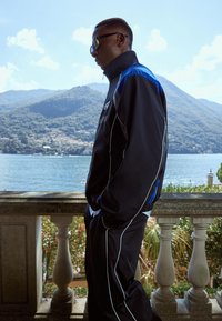 Czarny dres z niebieskimi akcentami i białym obszyciem, z wysokim kołnierzem i luźnym krojem. Noszone okulary przeciwsłoneczne, z malowniczym tłem.