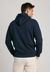 Felpa blu navy con cappuccio ampio; polsini e orlo a coste; materiale morbido; indossata sopra una maglietta bianca; vista posteriore semplice.