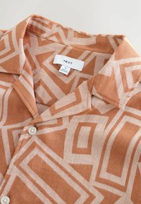 Chemise boutonnée avec grand motif géométrique beige et tan, étiquette de taille moyenne et col pointu.