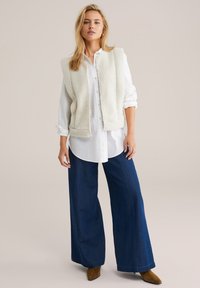 Bela teksturirana brezrokavnika preko dolge bele srajce z gumbi, v kombinaciji s širokimi temnodelnimi jeansom in rjavimi gležnjarji.