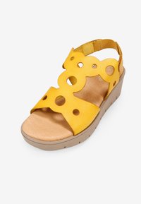 Sandalia de cuña de ante amarilla con patrones circulares cortados, correa para el tobillo ajustable y suela texturizada beige. Diseño de punta abierta.