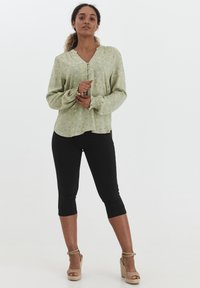 Blusa verde chiaro con scollo a V, maniche lunghe e motivo floreale; abbinata a pantaloni neri cropped e sandali con tacco a cuneo color sabbia.