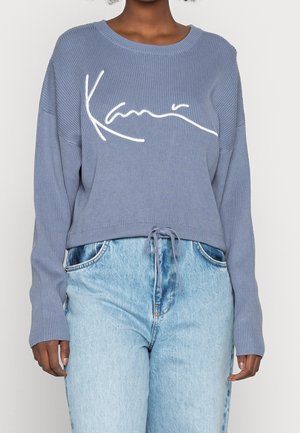 Personne portant un pull côtelé bleu clair avec le logo « Kani » en blanc et un jean taille haute bleu clair avec une taille à cordon.