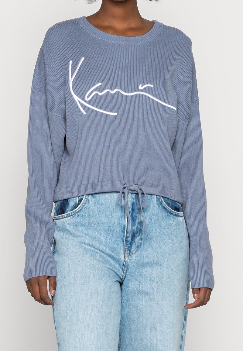 Personne portant un pull côtelé bleu clair avec le logo « Kani » en blanc et un jean taille haute bleu clair avec une taille à cordon.