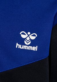 Tela azul con un acabado texturizado, que presenta un logo blanco de "hummel" y un diseño gráfico de una abeja estilizada, sobre un fondo negro.