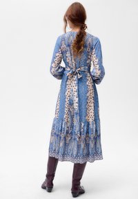 Blauwe en crèmekleurige jurk met patroon, lange mouwen, een strikdetail in de taille en een gelaagde zoom, gecombineerd met bordeauxrode laarzen tot kniehoogte.