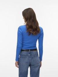 Woman with long brown hair wearing a bright blue long-sleeve top and high-waisted blue jeans, viewed from the back against white background.