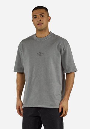 Mężczyzna ubrany w luźną szarą koszulkę z krótkim rękawem z małym czarnym napisem "PEGADOR" na środku klatki piersiowej oraz czarne spodnie.