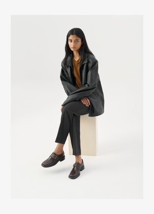 Schwarze lederne Oversize-Jacke, orangefarbenes Oberteil, graue maßgeschneiderte Hosen und braune Schnallenschuhe, sitzend auf einem hellen Block vor einem weißen Hintergrund.