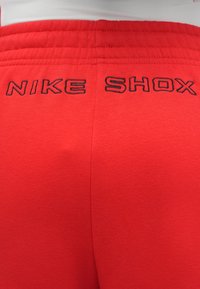 Κόκκινα αθλητικά σορτς από απαλό ύφασμα, με κεντητό μαύρο κείμενο "NIKE SHOX" κοντά στη μέση και άνετη, απαλή σχεδίαση χωρίς ραφές.