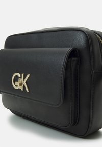 Borsa a tracolla in pelle nera con pattina frontale, accentata da logo dorato e chiusura con zip, caratterizzata da un design strutturato e una trama liscia.