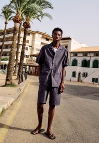 Jeune homme debout dans la rue portant une chemise grise à manches courtes et un short, des sandales noires, avec des palmiers et des bâtiments en arrière-plan.