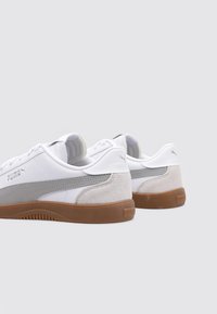 Zapatillas blancas con acentos grises, con una parte superior de piel suave y talón de ante, suela de goma marrón y logo de la marca en el lateral.