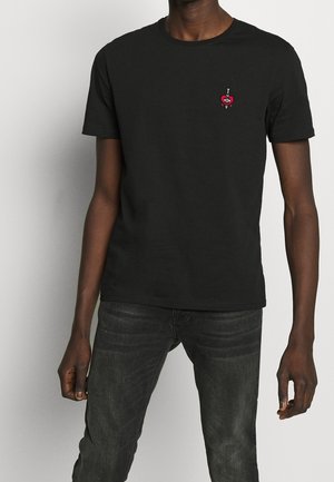 Mężczyzna w gładkiej, czarnej koszulce z krótkim rękawem z małym czerwonym sercem i grafiką sztyletu po lewej stronie klatki piersiowej, w połączeniu z ciemnymi dżinsami.