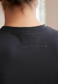 Černé sportovní tričko s hladkou texturou a plochými švy, s vyvýšeným logem "NEXT.STUDIO" na zadní straně poblíž výstřihu.
