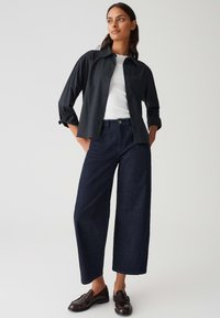 Donna in piedi con le mani in tasca, che indossa una camicia nera abbottonata sopra una maglietta bianca, jeans larghi scuri e mocassini scuri su uno sfondo semplice.