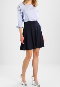 Marineblauer ausgestellter Rock aus weichem Stoff, kombiniert mit einem hellblau gestreiften Hemd. Das Model trägt silberne High Heels und steht vor einem neutralen Hintergrund.