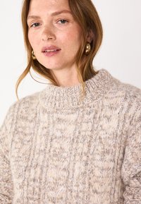 Maglione lavorato a maglia beige con un motivo testurizzato, caratterizzato da un colletto alto e una vestibilità ampia. Materiale caldo e morbido con dettagli di cucitura visibili.