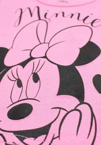 T-shirt en coton rose avec un design imprimé noir d'un personnage avec de grandes oreilles et un nœud, affichant des yeux et des mains expressifs.