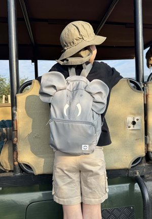 Harmaa norsun muotoinen reppu, jossa on suuret korvat, vetoketjutasku ja eläimen kasvokuvio. Lapsi käyttää sitä, hänellä on musta paita ja beiget shortsit.