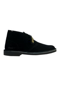 Botín chukka de ante negro con punta redondeada, cierre con cordones y suela de goma. Presenta una etiqueta dorada y un logotipo grabado en el lateral.
