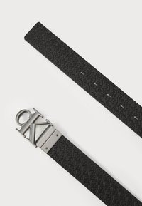 Zwarte gestructureerde riem met herhaald logo patroon, voorzien van een zilveren metalen gesp met gegraveerde letters. Duurzaam materiaal, slanke vorm, modern ontwerp.