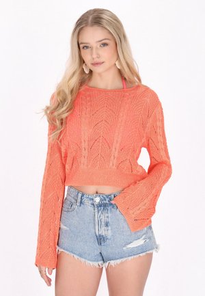 Blonde Frau mit lockeren Wellen trägt ein korallenfarbenes, langärmeliges Strick-Crop-Top und hoch taillierte, hellblaue Denim-Shorts mit Used-Details.