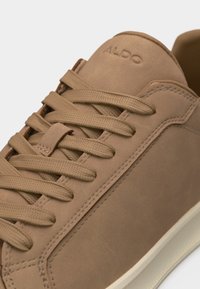 Gros plan sur une basket Aldo beige avec des lacets plats, des détails cousus sur la tige, et une semelle en caoutchouc blanche.