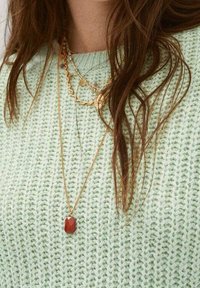 Pull vert clair en tricot avec un motif texturé ; colliers en or superposés avec un pendentif géométrique rouge. Les cheveux tombent en cascade sur l'épaule.