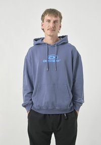 Kapuzensweatshirt in einem sanften, gedämpften Blau mit einer Brusttasche. Mit einem Logo in hellblau. Kombiniert mit locker sitzenden schwarzen Hosen.