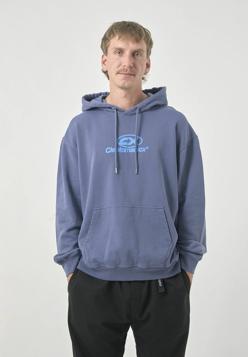 Kapuzensweatshirt in einem sanften, gedämpften Blau mit einer Brusttasche. Mit einem Logo in hellblau. Kombiniert mit locker sitzenden schwarzen Hosen.
