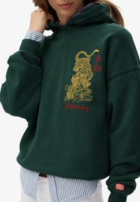 Hoodie vert avec un graphique de tigre jaune et le texte rouge "Superdry". Poignets et ourlet côtelés, porté par-dessus une chemise à col rayée bleue. Texture douce.