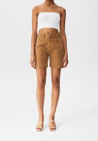 Shorts en denim marron avec un ourlet effiloché, taille haute et cinq poches. Assortis à un haut court blanc et des sandales blanches à lanières.