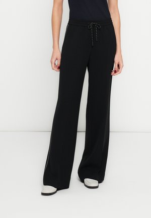 Personne portant un pantalon noir large à taille coulissée, un haut noir et des chaussures blanches, debout devant un fond blanc uni.