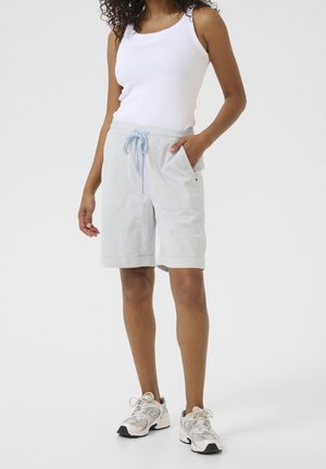 Donna con canotta bianca, pantaloncini al ginocchio a righe azzurri con coulisse e scarpe sportive bianche, in piedi contro uno sfondo semplice.