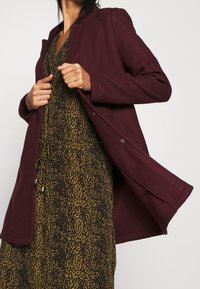 Bordeauxrode jas met een getailleerde kraag, een gladde textuur en drukknoopjes, gedragen over een bruine en gele luipaardprintjurk.