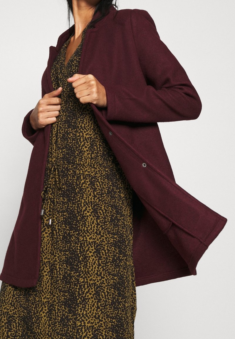 Bordeauxrode jas met een getailleerde kraag, een gladde textuur en drukknoopjes, gedragen over een bruine en gele luipaardprintjurk.