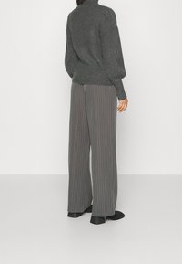 Person in grauen Nadelstreifen-Hose mit weitem Bein, dunkelgrauem geripptem Pullover und schwarzen Ankle Boots, steht vor einem schlichten weißen Hintergrund.