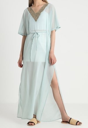 Mujer con un vestido largo azul claro transparente con escote en V dorado y cintura con lazo, combinado con sandalias planas abiertas en beige y negro.