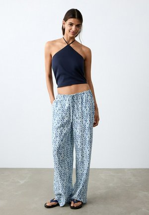 Femme portant un crop top halter bleu marine, un pantalon ample bleu clair à motifs et des tongs noires, debout les mains dans les poches sur un sol gris.