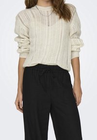 Lekki kremowy oversize'owy sweter z dzianiny z teksturowanymi pionowymi paskami i podwiniętymi rękawami, zestawiony z czarnymi spodniami ze sznurkiem w talii.