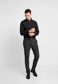 Camicia nera con bottoni abbinata a pantaloni grigi e scarpe nere. La camicia ha maniche lunghe e colletto classico, mentre i pantaloni hanno una vestibilità sartoriale.