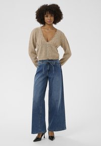Beige pletený kardigán s hlbokým výstrihom do V a gombíkovým zapínaním, kombinovaný s modrými džínmi s širokými nohavicami a čiernymi špicatými podpätkami. Hladká textúra, ležérny štýl.